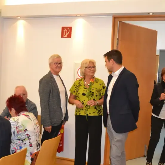 Mehrere Menschen stehen in einem Raum mit einem Feuerlöscher-Schild an der Wand. Eine Frau mit Brille hält eine Blume und spricht mit einem Mann. Die Frau auf der rechten Seite hält einen Glasbecher. Der Mann trägt einen Anzug und Lederschuhe.