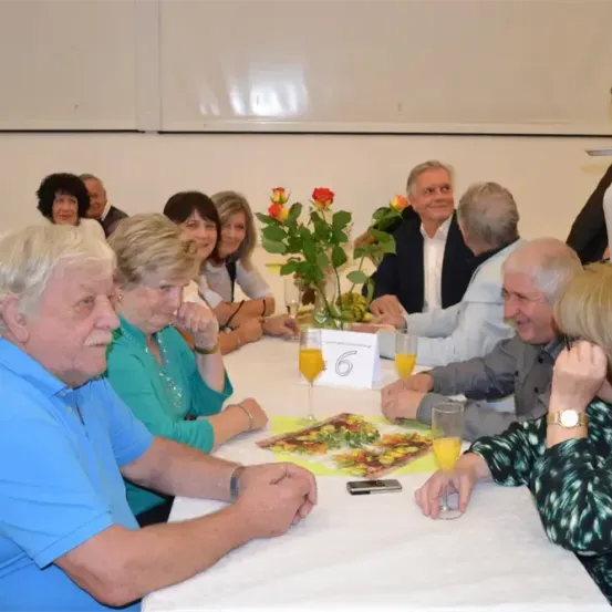Eine Gruppe älterer Menschen sitzt um einen Tisch herum, lächelt und plaudert. Ein Blumenstrauß und eine Sechs stehen auf dem Tisch. Einige von ihnen halten Gläser Orangensaft. Ein Mann steht hinter ihnen.