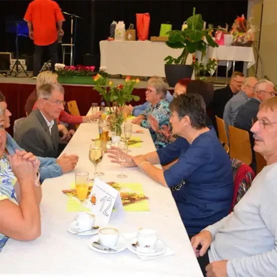 Eine Gruppe von Menschen sitzt an einem langen Tisch mit Tassen und Weingläsern, wahrscheinlich bei einer Veranstaltung. Im Hintergrund befinden sich Pflanzen und Blumen.