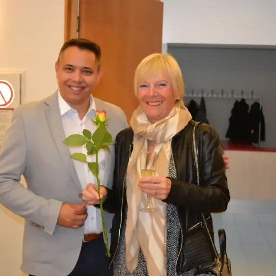 Ein Mann und eine Frau stehen nebeneinander und halten eine Rose und ein Weinglas, sie lächeln beide.