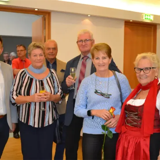 Eine Gruppe von Erwachsenen in formeller und halbformeller Kleidung steht eng beieinander und lächelt für ein Foto. Sie halten Weingläser in den Händen. Die Frau auf der rechten Seite trägt ein rotes Kleid und einen roten Schal. Hinter ihnen befindet sich eine Holztür.