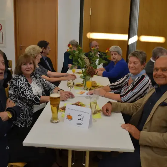 Eine Gruppe von Erwachsenen sitzt an einem Tisch mit Essen und Getränken. Sie lächeln und posieren für ein Foto.