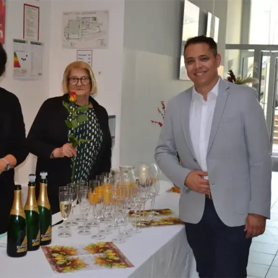 Drei Personen stehen hinter einem Tisch mit Champagnergläsern, Flaschen und einer Rose. Eine Frau hält eine Rose. Ein Mann lächelt und eine andere Frau schaut in die Kamera.