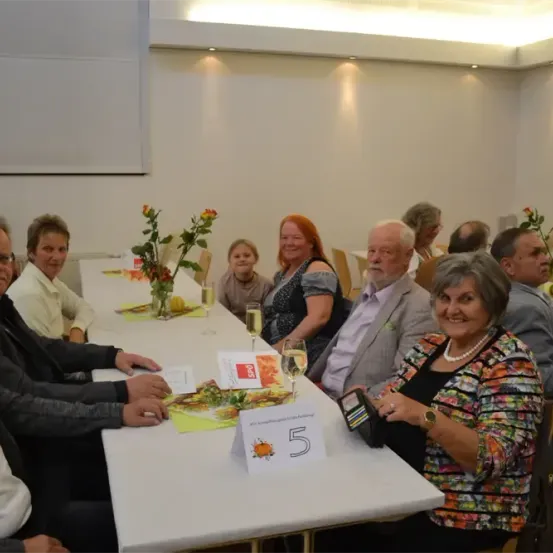 Eine Gruppe von Erwachsenen sitzt um einen langen Tisch mit der Nummer 5, Blumen und Weingläsern. Sie lächeln und scheinen eine Mahlzeit zu sich zu nehmen.