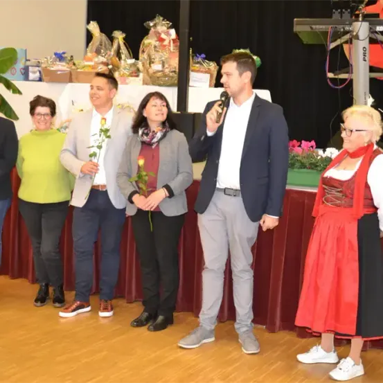 Eine Gruppe von Menschen auf der Bühne. Ein Mann spricht in ein Mikrofon, während andere Blumen halten. Ein Tisch mit verschiedenen Gegenständen steht hinter ihnen.