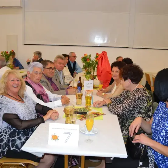 Eine Gruppe älterer Menschen sitzt um einen Tisch herum, wahrscheinlich bei einem Wiedersehen, mit Getränken und Blumen. Der Tisch hat eine weiße Tischdecke und eine Nummer 7.