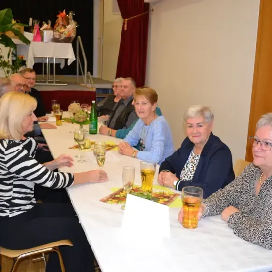 Mehrere ältere Frauen und Männer sitzen um einen langen Tisch herum, einige halten Gläser mit Bier in der Hand. Sie scheinen in ein Gespräch vertieft zu sein. Der Tisch ist mit Tellern, Gläsern und einer Flasche grünen Bieres besetzt. Dahinter befindet sich eine Bühne mit einem Tisch und einer Topfpflanze.