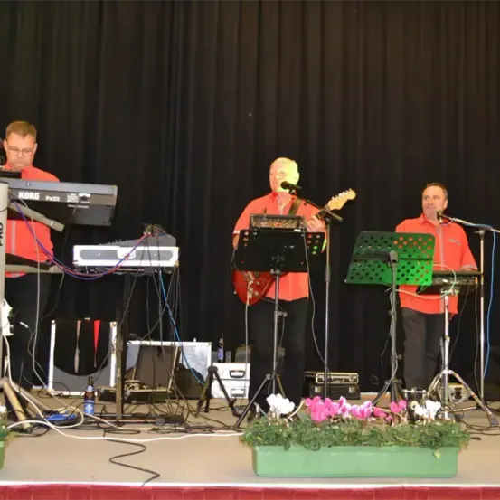 Drei Musiker stehen auf der Bühne. Der Mann auf der linken Seite spielt ein Keyboard, während der in der Mitte eine Gitarre spielt. Der Mann auf der rechten Seite singt in ein Mikrofon.