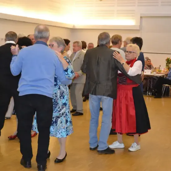 Mehrere ältere Paare tanzen auf einem Holzboden. Einige tragen traditionelle Kleidung. Im Hintergrund sind Tische mit Getränken und Essen aufgestellt.