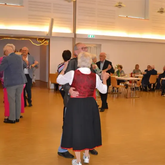Eine Gruppe älterer Erwachsener tanzt in einem großen Raum zusammen. Einige Leute sitzen an Tischen, und einige Tische haben Weingläser. Der Raum hat Holzböden und eine weiße Decke.