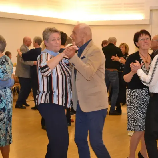 Mehrere ältere Paare tanzen in einem großen Raum. Sie tragen Freizeitkleidung und scheinen sich zu amüsieren. Der Raum hat Holzböden und weiße Wände.
