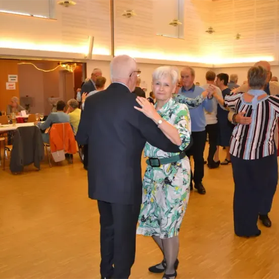 Ein älteres Paar tanzt, während andere ältere Menschen an Tischen in einem Saal sitzen.