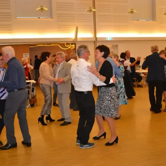 Mehrere ältere Erwachsene tanzen in einem Saal mit Holzböden. Ein Paar im Vordergrund umarmt sich, während andere in der Nähe tanzen.