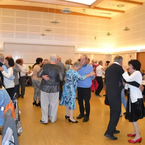 Mehrere ältere Paare tanzen in einem großen Saal, andere sitzen an Tischen. Der Saal hat Holzböden, weiße Wände und Lampen, die von der Decke hängen.