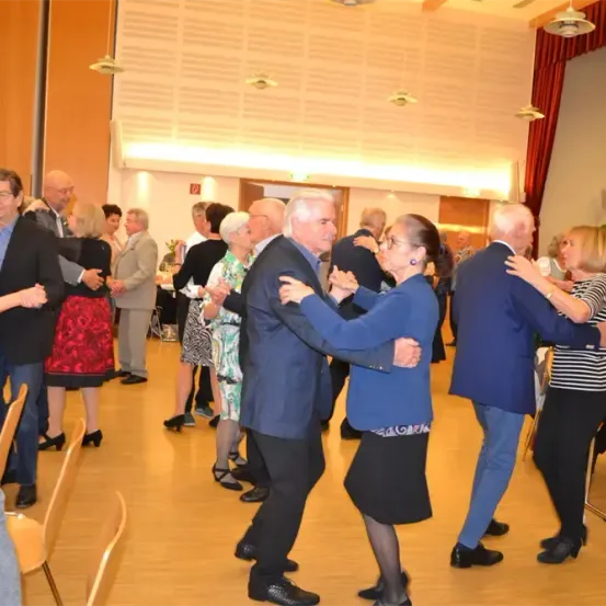 Eine Gruppe älterer Erwachsener tanzt in einem Raum zusammen. Sie sind in formeller Kleidung gekleidet und scheinen sich zu amüsieren. Der Raum hat Holzböden und mehrere Personen sitzen an Tischen.