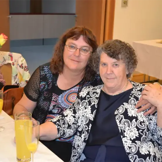 Zwei Frauen sitzen an einem Tisch mit Orangensaft und lächeln für ein Foto. Eine trägt eine Brille und ein gemustertes Oberteil, die andere eine schwarz-weiße Blumenjacke. Hinter ihnen ist eine Tür und eine Blumenarrangement zu sehen.