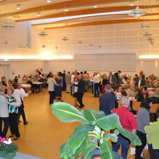 Ein großer Innensaal mit hoher Decke zeigt mehrere Personen an Tischen sitzend, einige stehend und tanzend, mit einer prominenten Pflanze im Vordergrund.