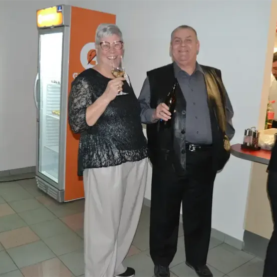 Ein älteres Paar steht eng beieinander, lächelt und hält Getränke. Die Frau hält ein Weinglas und der Mann eine Flasche. Sie befinden sich in einem Raum mit einem Kühlschrank und einer Theke mit einer Flasche. Ein anderer Mann steht in der Nähe.