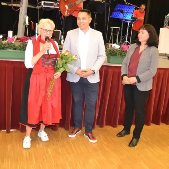 Eine Frau mit Brille und einem Blumenstrauß spricht in ein Mikrofon auf der Bühne. Ein Mann in einem weißen Blazer steht neben ihr und eine andere Frau in einem grauen Blazer steht neben ihm.