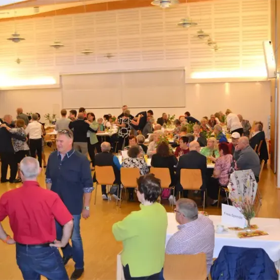 Eine Gruppe von Menschen versammelt sich in einem Saal, einige tanzen, andere sitzen. Der Raum ist mit Blumen dekoriert. Die Tische sind mit weißen Tischdecken bedeckt.