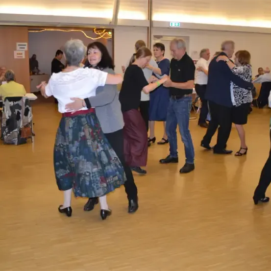 Mehrere ältere Erwachsene tanzen in einem Raum mit Holzböden. Sie tragen Freizeitkleidung und scheinen Spaß zu haben.