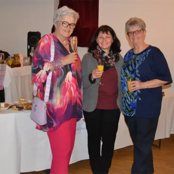Drei Frauen posieren mit Getränken bei einer Veranstaltung und stehen neben einem Tisch mit Essen. Dahinter befindet sich eine Wand mit einem Lautsprecher und einer Blumenarrangement.