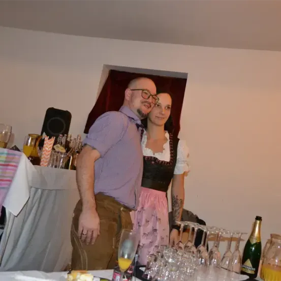 Ein Mann und eine Frau, lächelnd, stehen neben einem Tisch voller Weingläser, Flaschen und einem Lautsprecher. Sie tragen traditionelle deutsche Kleidung.