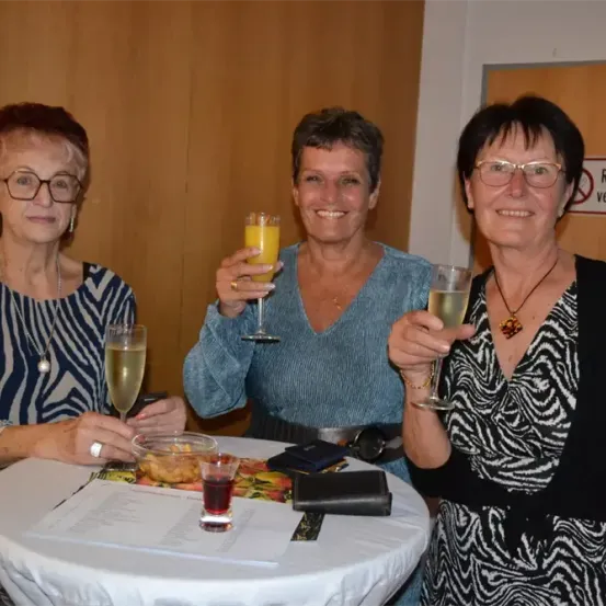 Drei Frauen stoßen mit Weinläsern an einem Tisch an, eine mit einem Ring, eine andere mit einer Halskette und eine dritte mit einem Handy. Ein Teller und eine Brieftasche sind auf dem Tisch, und ein Schild sagt Rauchverbot.