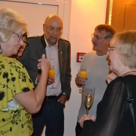 Eine Gruppe älterer Menschen steht und unterhält sich mit einander, dabei halten sie Gläser mit Orangensaft. Eine Person gestikuliert mit ihrer Hand.