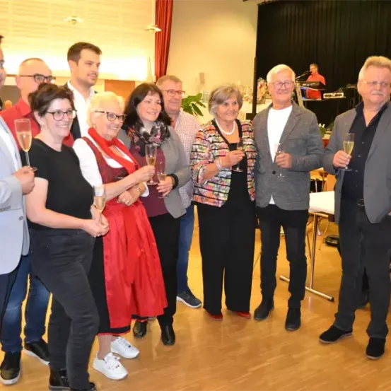 Eine Gruppe von Erwachsenen, einige mit Brille, in Freizeit- bis Halbformalkleidung, stehen zusammen und halten Weingläser. Der Hintergrund zeigt einen Raum mit Holzboden und einem schwarzen Vorhang.