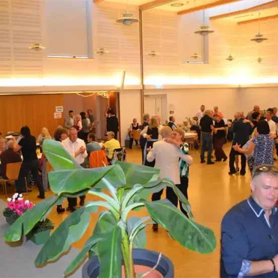 Eine Gruppe von Menschen tanzt in einem Saal mit Holzböden und Tischen, Stühlen und Topfpflanzen. Die Decke ist mit hängenden Lampen geschmückt.
