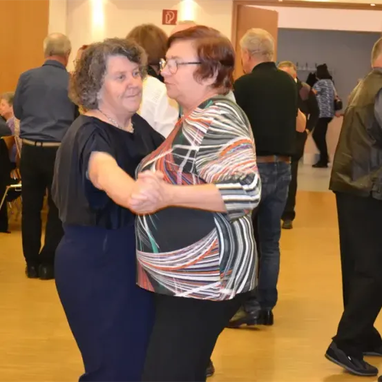 Zwei Frauen tanzen in einem Raum mit Holzböden, während andere stehen und zuschauen. Eine Frau trägt eine Brille, und eine andere trägt eine Halskette. Ein Schild mit dem Buchstaben P ist an der Wand sichtbar.
