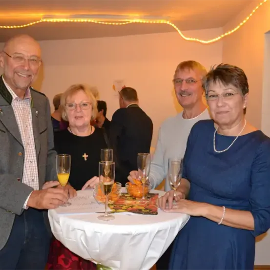 Vier Personen stehen um einen Tisch mit Weinglasern. Sie scheinen ein Ereignis zu feiern. Der Tisch hat eine weiße Tischdecke und einen dekorativen Teller.