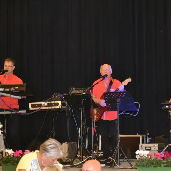 Eine Band tritt auf der Bühne auf, ein Gitarrist spielt Gitarre, eine Person spielt Keyboard und eine Person singt in ein Mikrofon. Eine Person steht vor einem Schlagzeug.