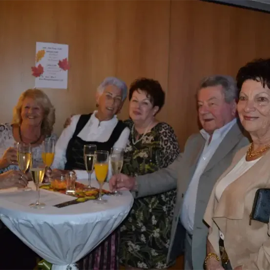 Eine Gruppe älterer Erwachsener versammelt sich an einem Tisch und hält Gläser mit Orangensaft. Sie lächeln und scheinen für ein Foto zu posieren. Dahinter eine Wand mit einem dekorativen Poster.