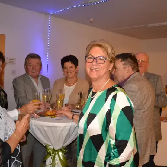 Eine Gruppe von Menschen versammelt sich bei einer gesellschaftlichen Veranstaltung, lächelt und hält Sektgläser in den Händen. Der Hintergrund zeigt eine Wand mit blauer Beleuchtung und einen Tisch mit Getränken und einer weißen Tischdecke.