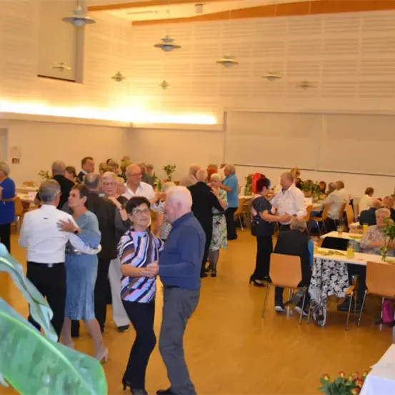 Eine Gruppe von Menschen tanzt und unterhält sich in einem Raum mit Tischen und Stühlen. Einige Menschen sitzen, während andere stehen. Es gibt Pflanzen und Lampen im Raum.