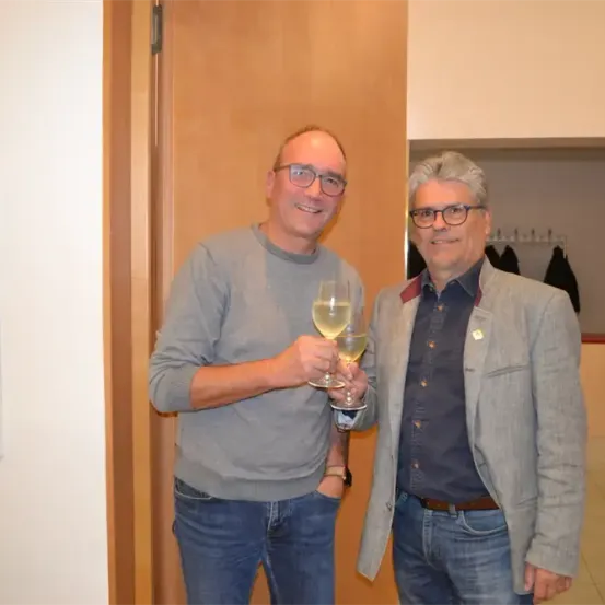 Zwei Männer, einer mit einem Weinglas in der Hand, stehen nebeneinander und lächeln, möglicherweise bei einer Feier. Der Mann auf der linken Seite trägt eine Brille und einen grauen Pullover, während der Mann auf der rechten Seite einen grauen Blazer und ein blaues Hemd trägt.