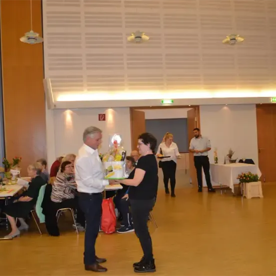 Eine Gruppe von Menschen versammelt sich in einem Saal, einige sitzen an Tischen, andere stehen. Ein Mann in einem weißen Hemd überreicht einer Frau in Schwarz einen Kuchen. Im Hintergrund gehen zwei Frauen zu einem Tisch.