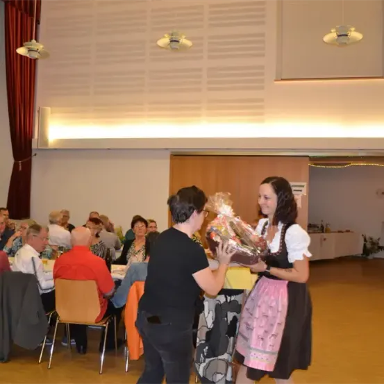 In einem Raum übergibt ein Mann einen Geschenkkorb an eine Frau in traditioneller Kleidung. Zuschauer sitzen hinter ihnen an Tischen, und im Hintergrund stehen Topfpflanzen.