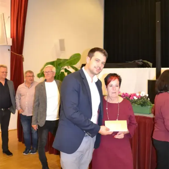 Ein Mann in einem blauen Anzug überreicht einer Frau in einem weinroten Kleid eine Auszeichnung, während mehrere Personen hinter ihnen stehen.