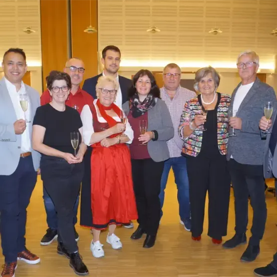 Eine Gruppe von Menschen steht in einem Raum, lächelt und hält Weingläser. Sie scheinen ein Ereignis zu feiern.