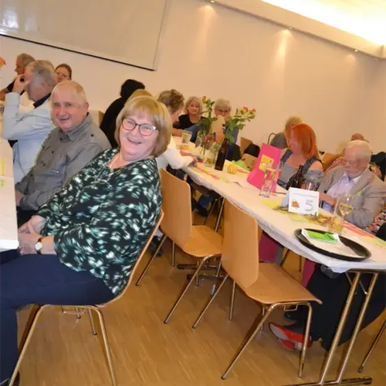 Eine Gruppe älterer Menschen sitzt an einem langen Tisch, lächelt und plaudert. Der Tisch ist mit Blumen und Tellern dekoriert. Sie befinden sich in einem Raum mit Holzböden und weißen Wänden.