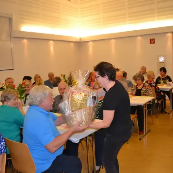 Eine Indoor-Veranstaltung mit einer Gruppe älterer Erwachsener, eine Frau präsentiert einen Korb einem sitzenden Mann. Andere Teilnehmer sitzen an Tischen, und einige stehen. Der Raum ist gut beleuchtet.