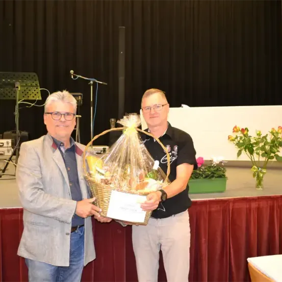 Zwei Männer halten einen Korb mit verschiedenen Lebensmitteln auf der Bühne. Hinter ihnen steht ein roter Vorhang, und ein Mikrofon ist auf einem Ständer aufgebaut.