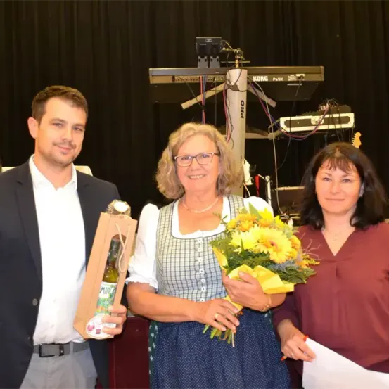 Drei Personen stehen vor einem schwarzen Vorhang. Ein Mann im Anzug hält eine Flasche und lächelt. Die Frau im karierten Kleid hält einen Blumenstrauß. Die Frau im braunen Hemd hält ein Stück Papier. Hinter ihnen steht ein Notenständer mit einem Korg-Keyboard.