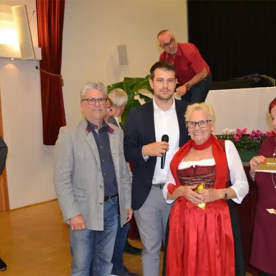 Eine Gruppe von Menschen steht in einem Innensaal. Der Mann mit dem Mikrofon scheint eine Rede zu halten. Eine Frau in einem roten Kleid hält einen Blumenstrauß. Ein Mann steht hinter ihnen, und eine Topfpflanze steht in der Nähe der Wand.