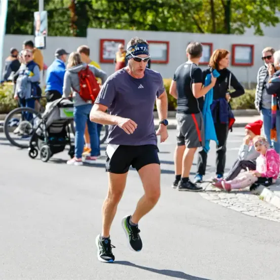 Ein Mann läuft in einem Marathon, während mehrere Menschen herumstehen und zusehen. Er trägt ein lila Adidas-Shirt, eine Sonnenbrille und schwarze Shorts.