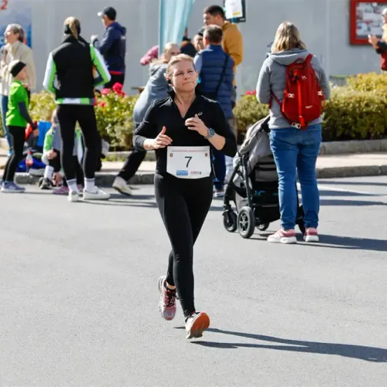 Eine Frau in einem schwarzen Outfit mit der Nummer 7 auf dem Shirt läuft bei einem Marathon. Dahinter stehen einige Leute und beobachten.
