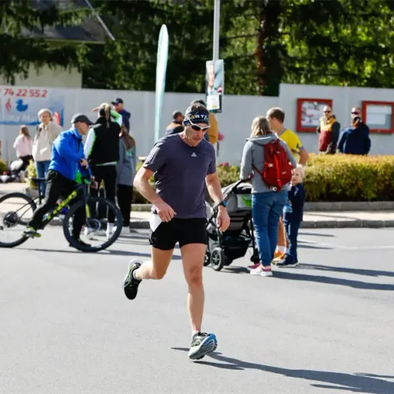 Ein Mann joggt auf der Straße, während Menschen herumstehen und eine Frau einen Kinderwagen schiebt. Ein Fahrrad ist in der Nähe geparkt.